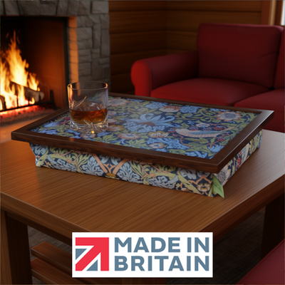 The William Morris Strawberry Thief lap tray with wooden frame, shown in a cosy fireside setting with a glass of whisky, alongside the Made in Britain logo.